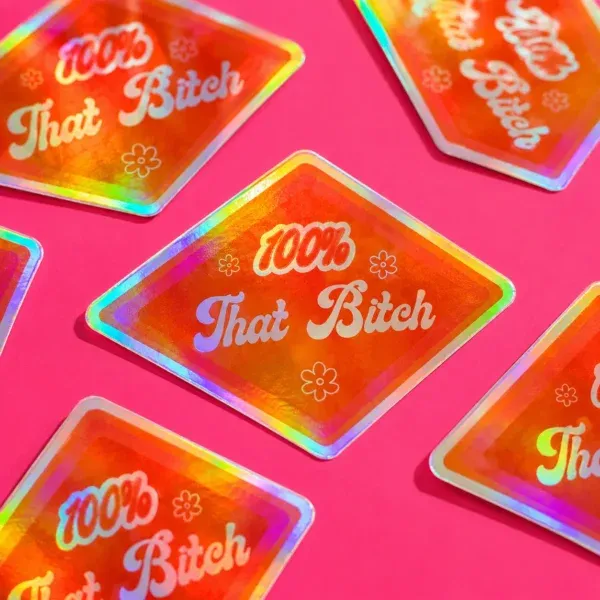 100% That Bitch Holographic Sticker on hot pink table.